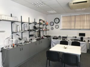 Photo shows the left side of our Hanley Resource Centre. On the left wall are cupboards and shelves displaying a variety of equipment including lamps, magnifiers, eye shields, games and labeling equipment, in the back left corner are talking book players and accessible radios. On the back wall are tables with white table cloths displaying video magnifiers of various sizes and on the actual wall are large print wall clocks in a variety of sizes. In the centre of the room are 2 white tables with 4 black chairs around them. Out of shot to the right are more displays or other types of equipment including mobility aids and kitchen equipment. 