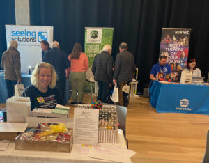 Photo taken at our 2023 Information Day at the Catalyst Building at Staffordshire University. In the foreground our trustee Sandra sits smiling at a table, on the table in front of her are a variety of raffle prizes including hampers and tickets for days out. Behind her across the back wall are exhibitors with their logos displayed on their roller banners. From left to right are Seeing Solutions, Staffordshire Cricket and the New Vic Theatres/ Appetite. We can see the backs of attendees facing the exhibitors, talking to them, trying equipment and collecting information.