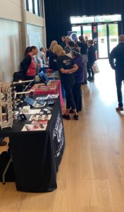 Photo taken at our 2023 Information Day at the Catalyst Building at Staffordshire University. On the left of the photo are exhibitor tables including Associated Optical and the RNIB but more tables can be seen going into the distance of the shot. They all have different coloured table cloths and a variety of information leaflets and equipment on their tables. Lots of attendees can be seen talking to the exhibitors.