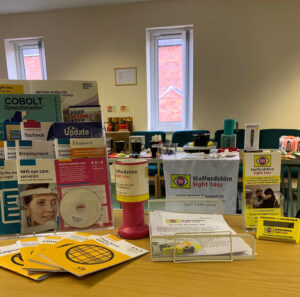 This photo was taken at our monthly Leek drop-in at the Salvation Army Building on Salisbury Street. In the foreground are tables that have a variety of information leaflets, newsletters and signature guides on the them as well as a pink charity collection box. In the background more tables can be seen with lots of different equipment including talking kitchen and labelling equipment. Behind that is a wall and window.