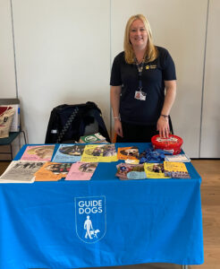 This photo shows one of our exhibitors Wendy from Guide Dogs at her stand, she stands behind the table smiling. On the table is a blue tablecloth with the Guide Dogs logo on the front. On top of the table are a variety of multi-coloured information leaflets. Wendy wears a navy blue polo shirt with the Guide Dog logo in yellow. Also on the table is a box of celebration chocolates.