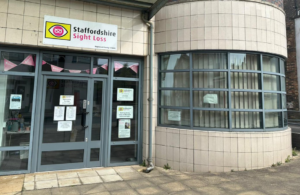 This photo shows the outside of our Hanley Resource Centre which is open from 10am til 4pm every Tuesday. To the left is a glass door surrounded by windows, in the windows are signs and posters. Above the door is a Staffordshire Sight Loss Association sign with our logo in the colours pink, yellow, black and white. To the right is a large bay window, the building is covered in cream coloured tiles.
