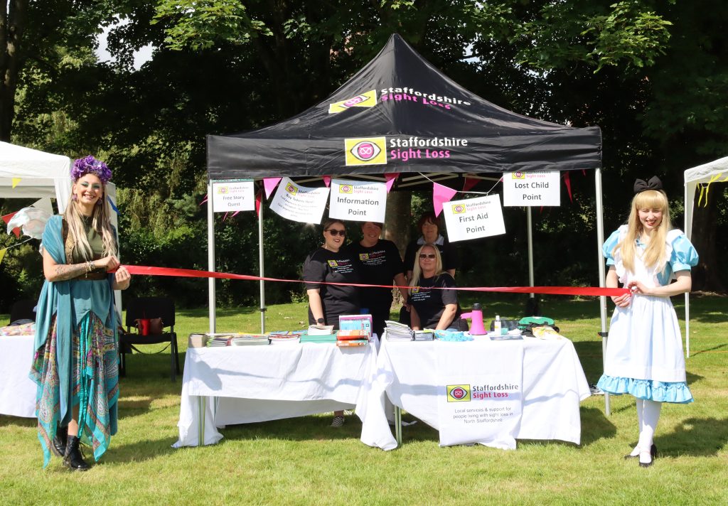 This image shows a black marquee with the Staffs sight loss association logo on the roof, under the marquee 4 volunteers are visible, in front of the marquee there a 2 ladies on the right is a lady dressed up as alice in wonderland and on the right is a lady dressed as a fairy, they are both holding a red ribbon ready for it to be cut to start the community fun day