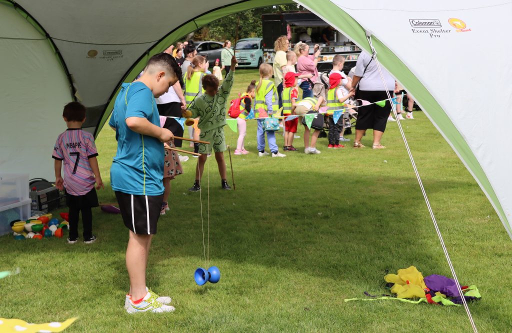 Image shows a young boy under a marquee playing with a blue diablo, he is concentrating hard on it. In the background there are other children looking at what is going on