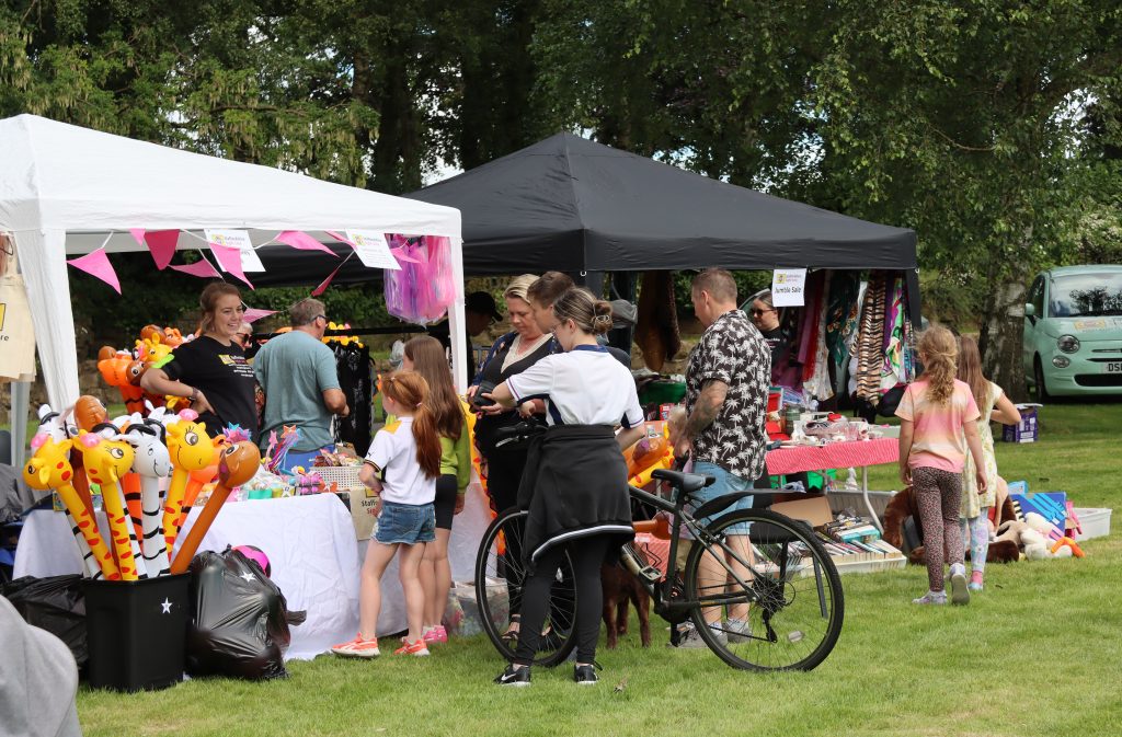 Image shows 2 marquees, new white and one black. under the white marquee we can see Beth in her SSLA tshirt selling pocket money toys, Beth looks happy as she talks to some children. Toys for sale include large inflatable animal sticks, fairy wings and wands and dinosaur eggs. Under the black marquee there are a range of second hand items for sale. In front of both marquees there is a group of people, both children and adults, including one man who is holding a bike.