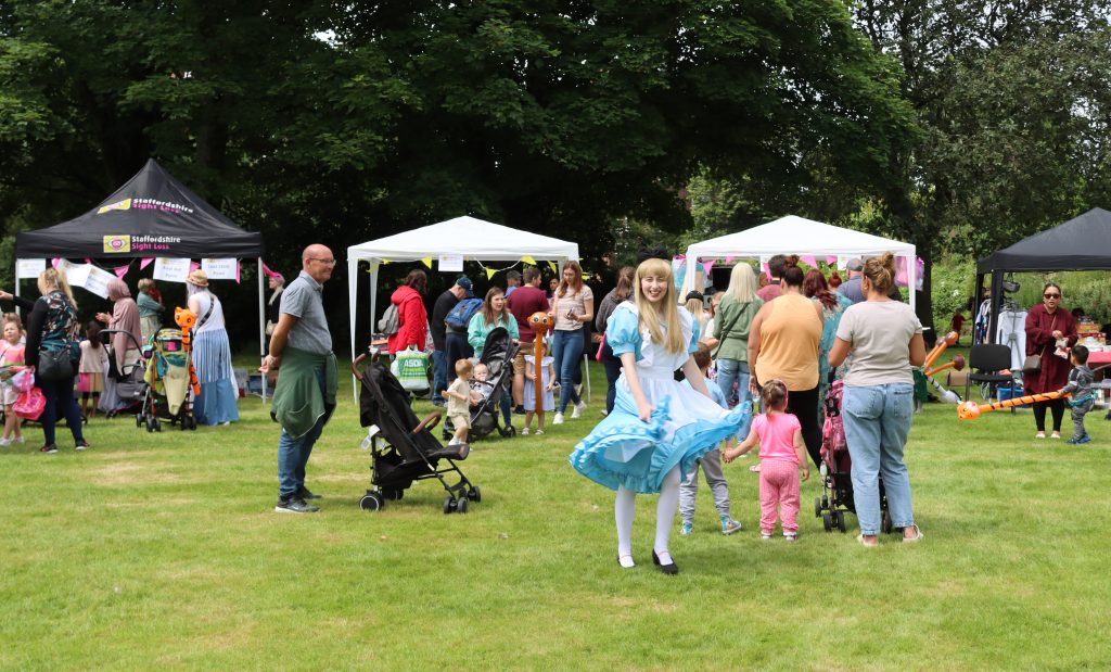 The image shows a lively outdoor event taking place on a grassy area. There are several people gathered, including adults and children. In the foreground, a person dressed in a blue and white costume, resembling Alice from "Alice in Wonderland," is interacting with the crowd. Nearby, a child in a pink outfit is holding hands. In the background, there are white activity tents with people standing around them. The scene is set against a backdrop of trees