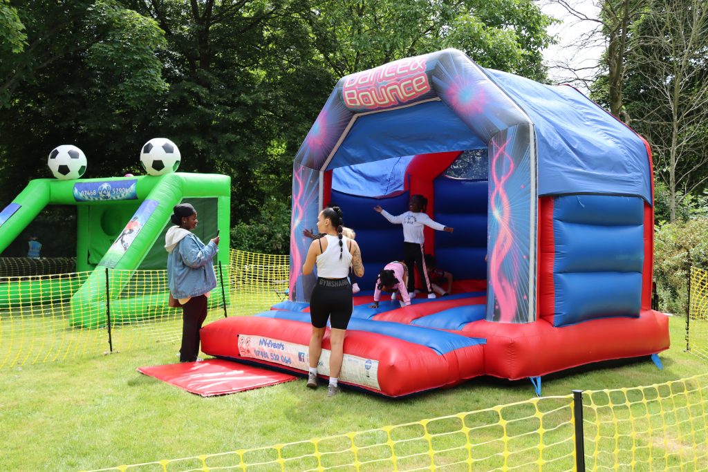 Image shows a blue and red bouncy castle with three children bouncing on it while some adults stand watching them. The children look like they are having fun. In the background an inflatable penalty shoot out is also visible.