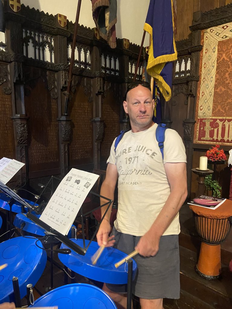 This photo shows Gareth standing in front of a blue steel drum, he is tapping his drumsticks onto the surface of the drum and a music stand and sheet of music can be seen facing away from him. He is wearing a cream-coloured t-shirt, grey shorts, and sunglasses on his head. In the background, decorative wooden panels cover the walls of Stoke Minster and a blue and yellow flag is hanging from a flag pole. There are also red flowers, candles, and another drum in the background.