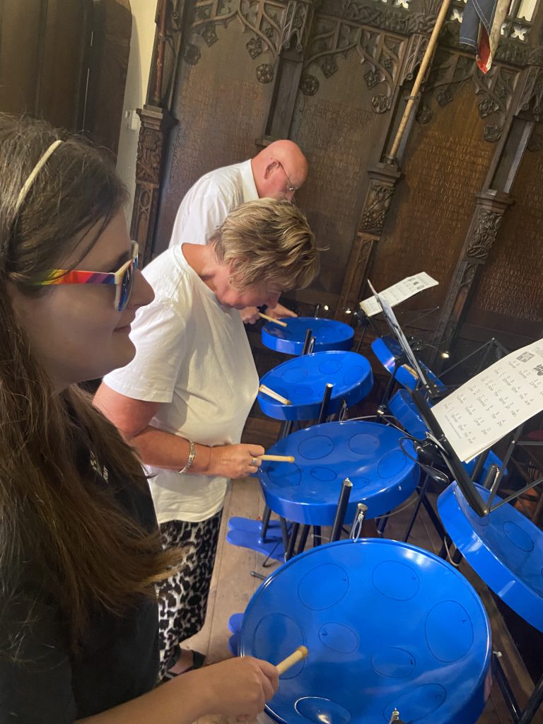 This image shows several people playing blue steel drum instruments in what appears to be a church or similar ornate building. In the foreground, Klaudia can be seen with long dark hair wearing sunglasses. Behind her is another person with blonde hair and wearing a leopard print skirt holding drumsticks over one of the blue steel drums also playing a steel drum. Further back, there's a man in the background. The steel drums are a bright blue colour and appear to be arranged in a semicircle. There are music stands visible with sheet music on them. The background shows intricate wooden paneling or screens with Gothic-style carvings, typical of church architecture. This setting adds an interesting contrast between the traditional surroundings and the more modern instruments.