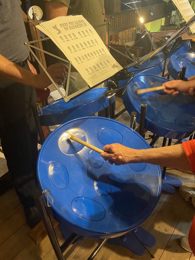 This image shows a close-up view of people playing steel drums, also known as steel pans. The main focus is on a bright blue steel drum in the foreground, with a person's hand visible holding a drumstick and striking one of the note areas on the drum's surface. Behind this drum, you can see parts of other blue steel drums arranged together. There's a music stand holding sheet music, which appears to be a chart or guide for playing the steel drums. The title on the sheet music is partially visible but not entirely clear in the image. In the background, you can see some other musical equipment and what looks like a rehearsal or performance space. The lighting suggests an indoor setting. The image captures a moment of music-making, and the hands-on process of playing it.