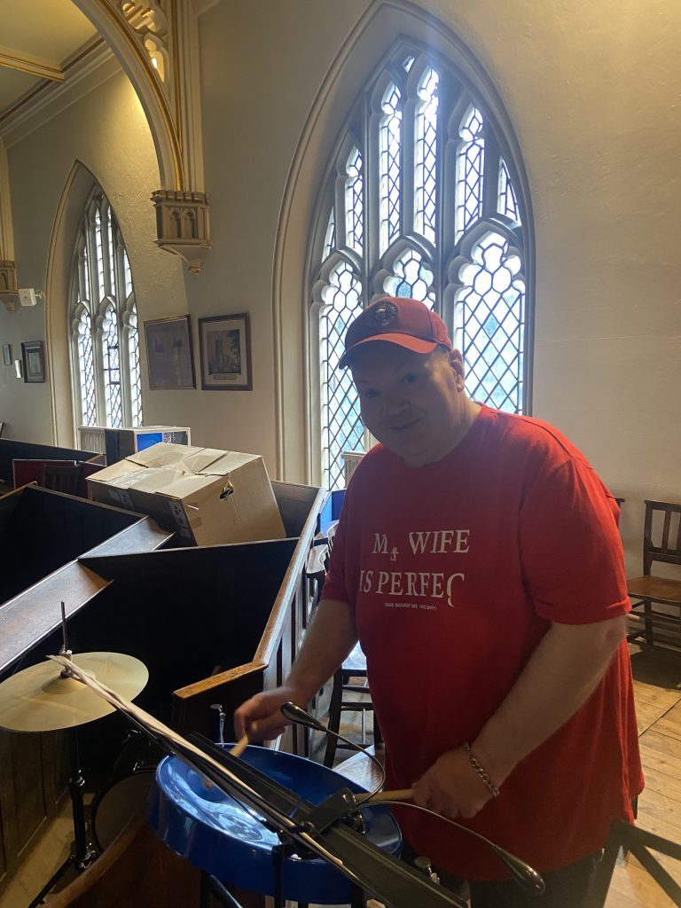 This image shows someone inside a church. He is standing next to some musical equipment, specifically a drum set. They are wearing a red t-shirt. He's also wearing a baseball cap. The setting is quite distinctive, with large Gothic-style arched windows visible behind him. These windows have intricate tracery and leaded glass designs typical of church architecture. The interior walls are light-coloured, and there are some framed pictures or artworks visible on the wall. He is smiling at the camera and appears to be in the process of setting up or adjusting the drum set. There are also some cardboard boxes visible. The architectural elements, including the ornate arched doorways and windows, strongly indicate this is taking place in a church, though it's being used in an unconventional way with the presence of modern musical equipment.