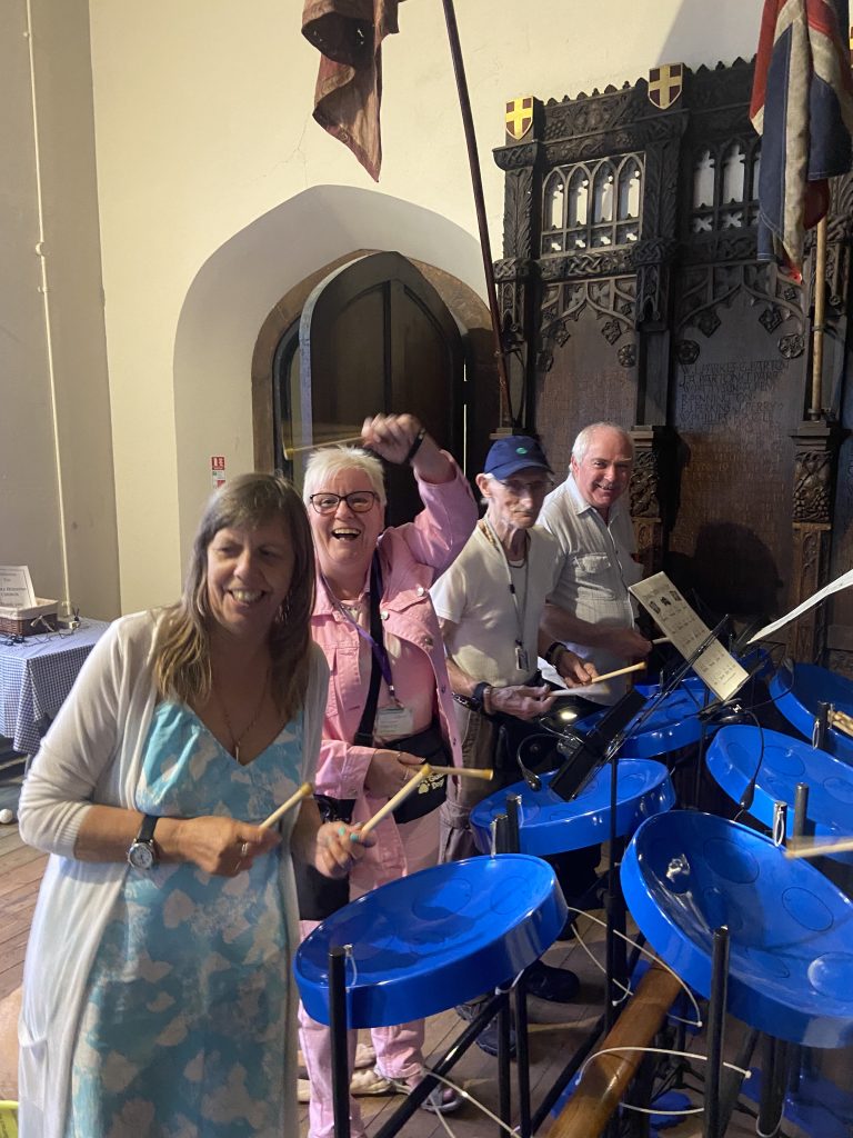 This photo shows Angela, Hazel, John, and Geoff standing next to the Steel drums that they have been playing, each holding a pair of drumsticks. They are smiling and Hazel is laughing and holding one of her drumsticks in the air above Angela’s head.