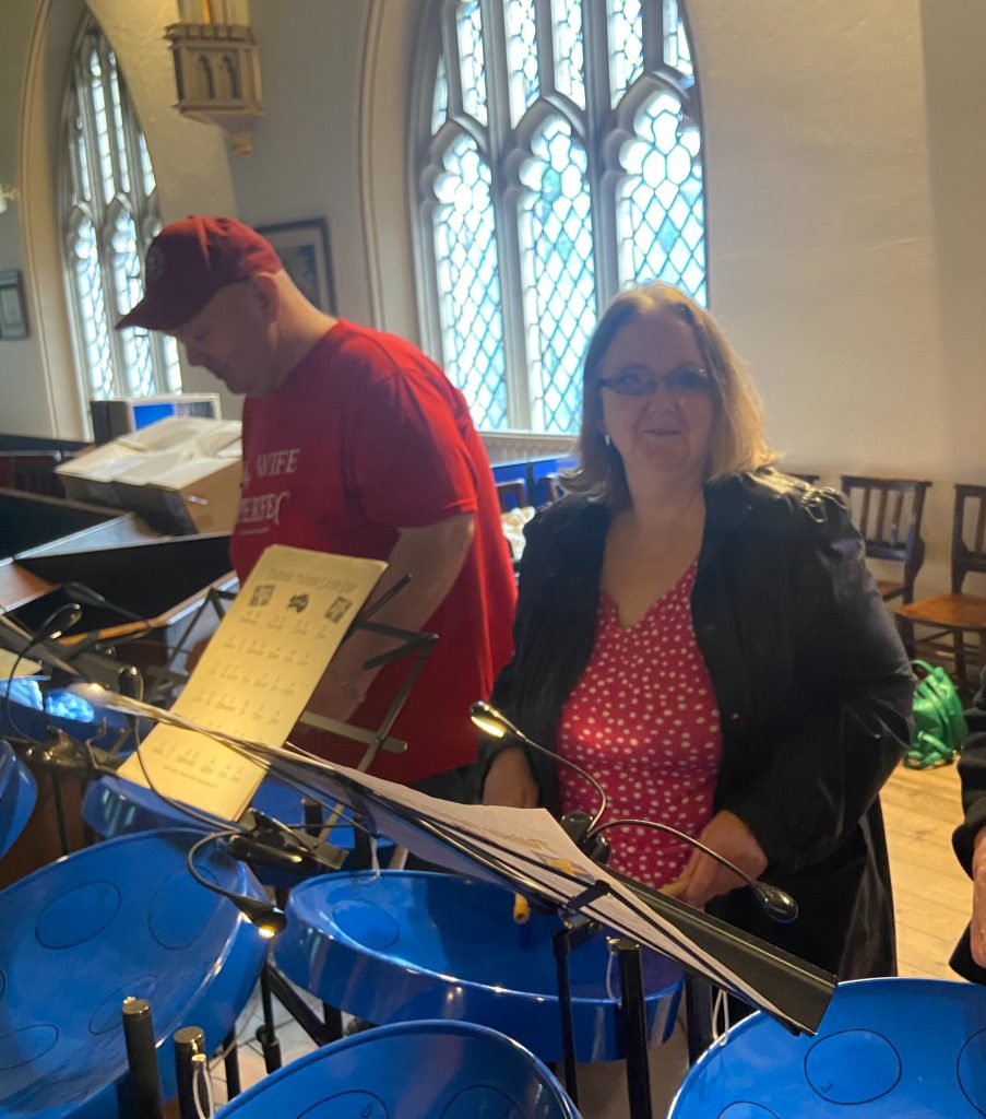 This image was taken inside a church, as evidenced by the ornate stained glass windows visible in the background. The room has high ceilings and Gothic-style architecture. In the foreground, there are two people standing near what looks like a set of steel drums or similar percussion instruments. These instruments are a bright blue color. On the left is a someone wearing a red baseball cap and a red t-shirt. On the right is a woman with shoulder-length blonde hair, wearing glasses. She's dressed in a black jacket over a red and white polka dot top. Both individuals appear to be looking at or working with sheet music on stands in front of them, suggesting they might be preparing for a musical performance or practice session. The setting creates an interesting contrast between the traditional church architecture and the modern steel drum instruments.