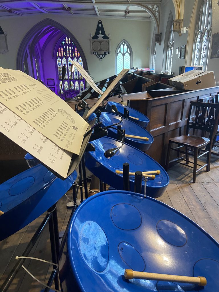 This photo shows the Steel Drums at Stoke Minster, the picture shows 4 drums in a row starting in the foreground and going into the distance, the drums are royal blue colour and each one is shaped like a very shallow bowl. On each drum's surface, circular shapes are drawn to show where to hit the drum to achieve each note. The notes from A to G are written on each of these shapes. Each drum has a pair of drumsticks placed on it. To the left of these drums are another 4 drums facing them, mostly out of shot. A music stand with music sheets can also be seen. The Steel drums are located upstairs in Stoke Minster, and colourful stained glass windows are in the background.