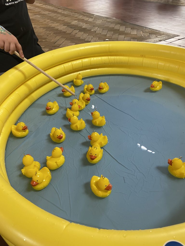 This is a photo of our hook-a-duck pool, it is a yellow inflatable pool with a blue base and 20 yellow ducks floating in it with hooks on their heads. To the left a hand can be seen holding a hook-a-duck rod as someone tries to hook a duck!