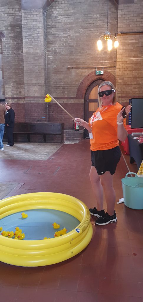 This photo shows a lady from the MS Society stall at the event, she is wearing an orange MS Society t-shirt, black shorts, and black trainers. She is also wearing a pair of simulation glasses. In her hand is a hook-a-duck rod and she has successfully caught a duck on the end. At her feet is the hook-a-duck pool which is a yellow inflatable pool with a blue base and lots of yellow ducks floating in it.