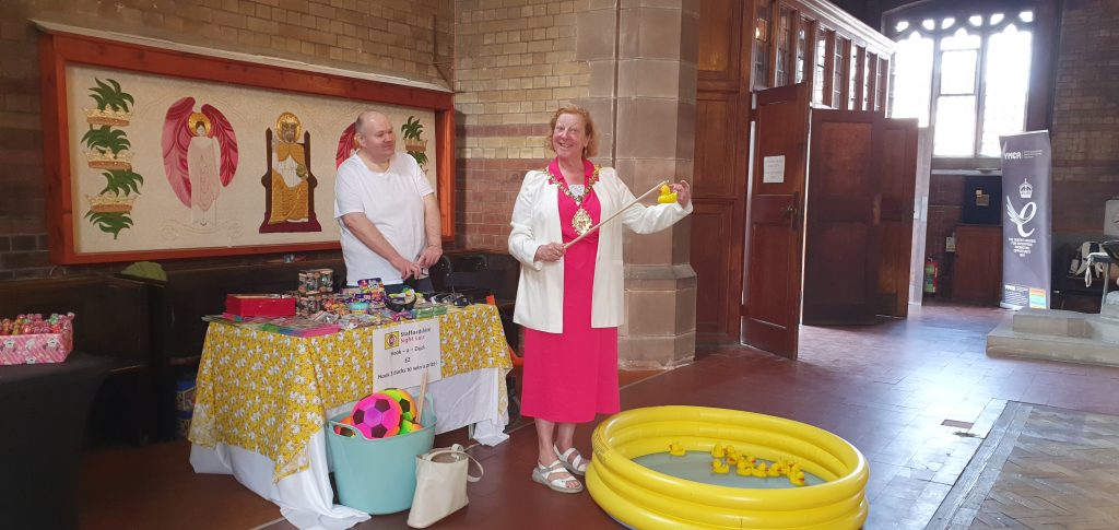 Photo taken at the Hanley, Bucknall and Bentilee PCN fun day at All Saints Church, Joiner’s Square. The SSLA team attended to raise awareness and to fundraise with a hook-a-duck game. This photo shows Trustee Ian Knowles standing behind a table with a yellow tablecloth covered in pictures of ducks. In front of the table stands the Lord Mayor of Stoke-on-Trent Councillor Lyn Sharpe wearing a pink dress, white jacket, and her gold Mayoral Chains. At her feet is an inflatable yellow paddling pool with a blue base, inside the pool float 20 yellow ducks with hooks on their heads. Lyn has a wooden stick with a hook at the end in her hand and on the end of the hook she has successfully caught a duck! She is smiling at the camera. On the table are a variety of prizes (mostly toys such as slinkies and games). In the background are the stone walls of the church, a wooden clad wall with doors, and decorative artwork. Ian is looking at Lyn and smiling.