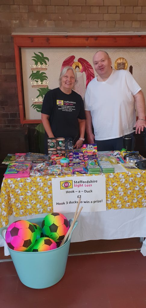 This photo shows our Administrator Tricia and Trustee Ian standing behind a table full of prizes for our hook-a-duck game. The table is covered with a white tablecloth overlaid with a yellow tablecloth covered in pictures of ducks. A sign at the front says ‘Staffordshire Sight Loss, Hook-a-duck £2, hook 3 ducks to win a prize!’. Prizes on the table include fun sand, dinosaur eggs, sticker books, and slinkies, in front of the table is a large bucket with neon rainbow footballs and hook-a-duck rods. Also on the table are SSLA leaflets and a variety of sim specs that simulate various eye conditions.