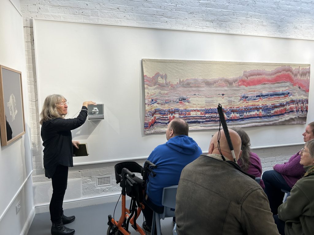 This image shows a woman with gray hair presenting to a small group of seated attendees. She's gesturing towards a small display on the wall while a large abstract painting hangs behind her.