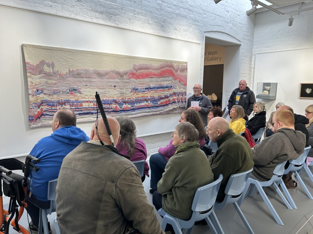 This image provides a wider view of the gallery space. The same large abstract painting is visible, and more attendees are seated facing it. The space has white brick walls and track lighting. A sign for the "Jim Wain Courtyard Gallery" is visible.