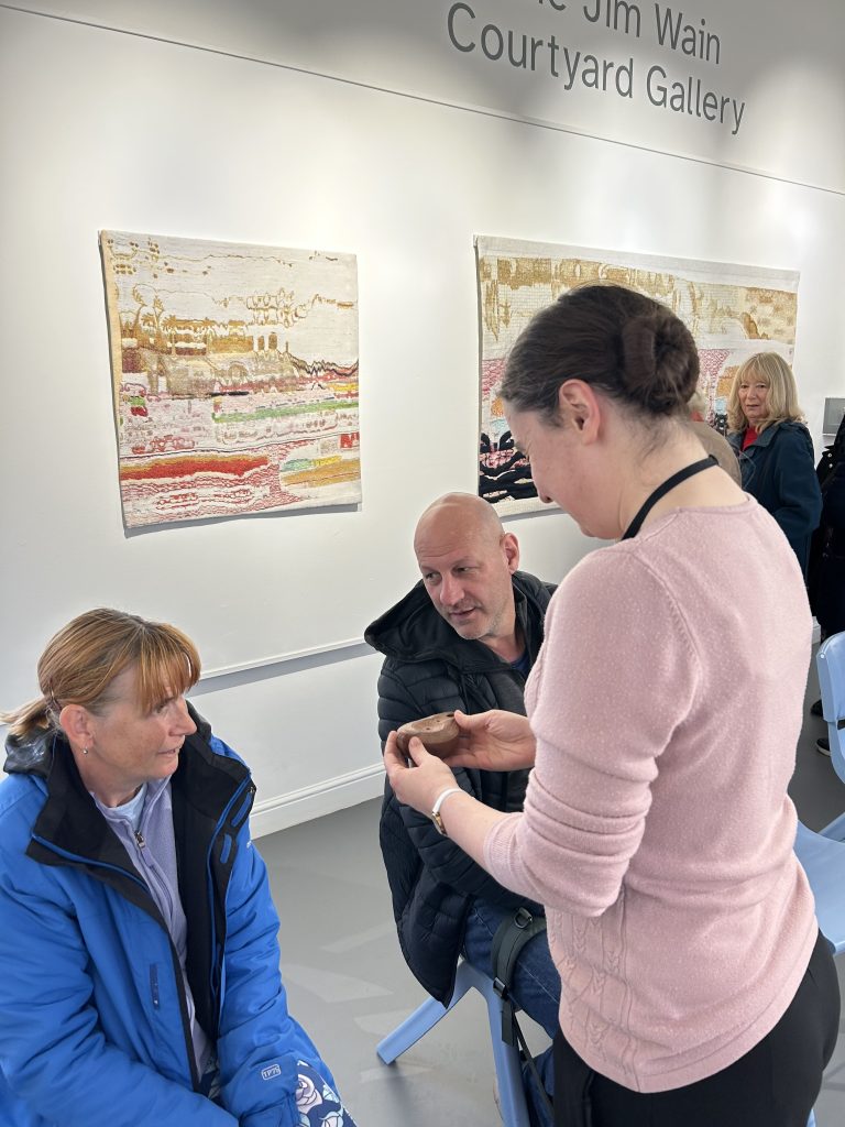 This image shows a closer view of some artworks on display. They appear to be textile or mixed media pieces with abstract designs in earth tones. A few attendees are examining the art, including someone holding what looks like a tactile object for a hands-on experience of the artwork.