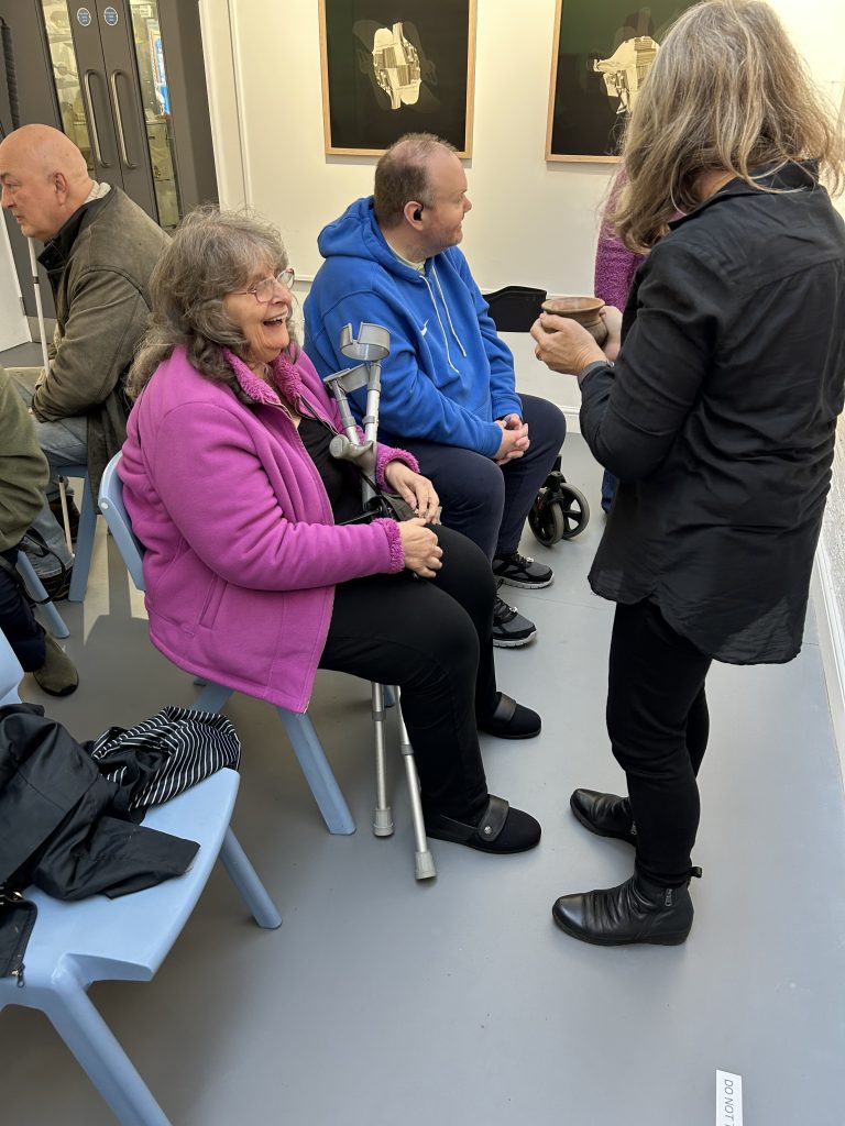 This image captures a small group interacting. A woman in a bright pink jacket is smiling, seated next to a man in a blue hoodie. Another person is showing them something small they're holding, possibly a tactile representation of artwork for a hands-on experience. Framed artworks are visible on the wall behind them.