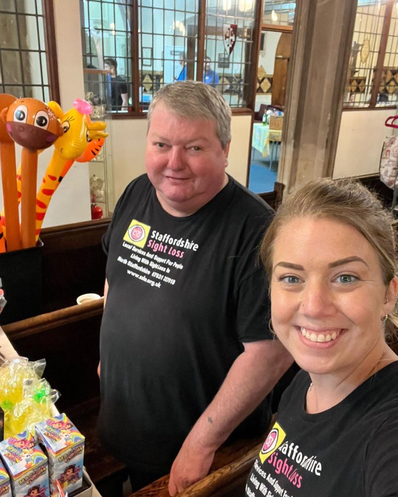 The photo shows two people standing indoors, at an event. They are both wearing black T-shirts with the logo and text for “Staffordshire Sight Loss.” Ian is smiling slightly. Beth is smiling broadly. In front of them, there are various colorful items on display, including inflatable toys shaped like animals and boxes with bright designs. The background features large windows with a grid pattern and some people and tables visible through the glass.