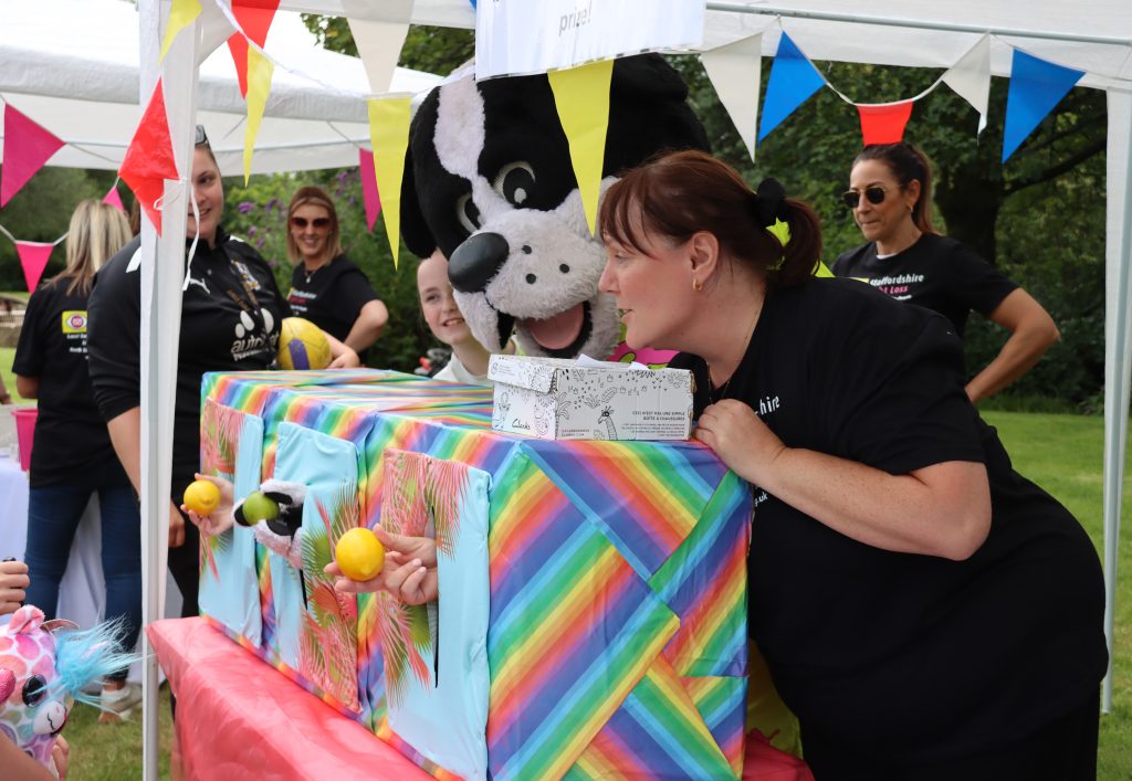 This image shows boomer, the port vale mascot, who is a large black and white dog taking part in the human fruit machine. He and 2 other volunteers have their hand through a large rainbow striped box and are holding a piece for fruit each (2 lemons and an apple)