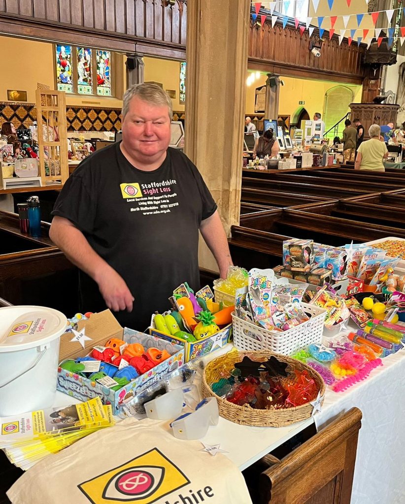 The image shows Ian standing behind a table filled with various colorful items, likely for sale. He is wearing a black T-shirt with the logo and text for “Staffordshire Sight Loss”. The table is covered with a white cloth and has a variety of small toys, candies, and other items displayed in baskets and containers. The setting is a church; there are wooden pews, stained glass windows, and a high ceiling with bunting decorations hanging across. Other people are visible in the background, some browsing other tables.