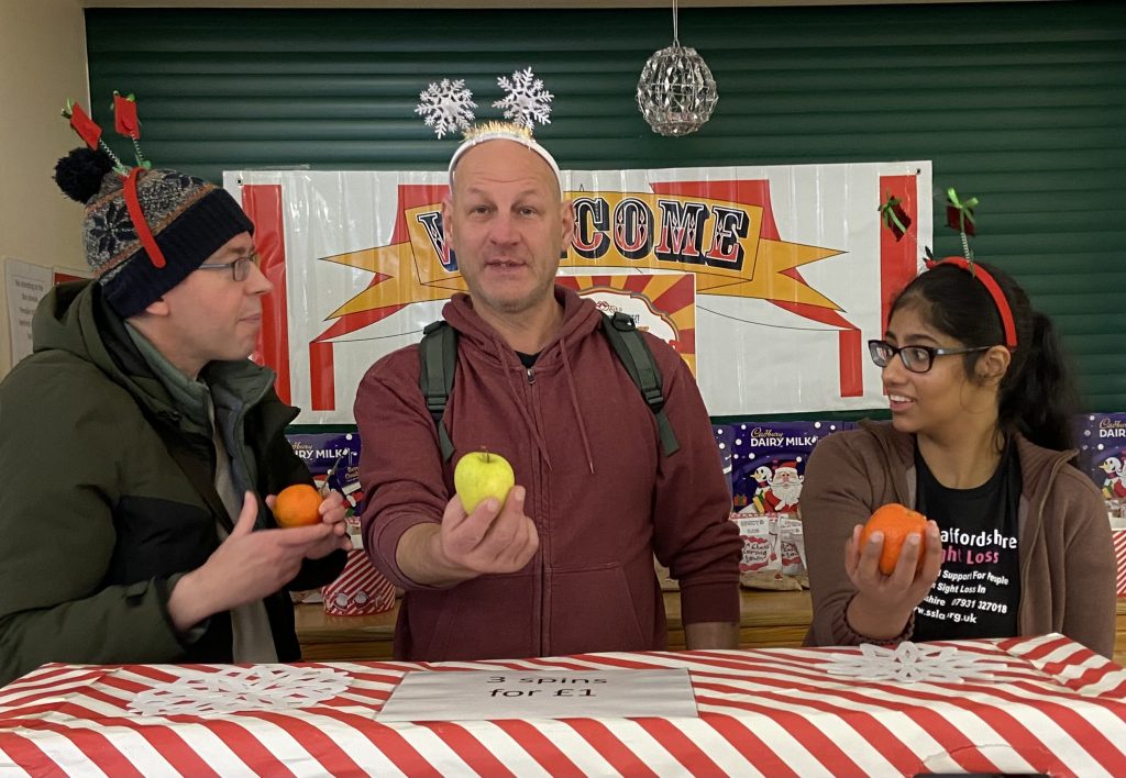 This image shows three people standing behind a red-and-white striped table with a sign that says "3 spins for £1." Behind them is a "Welcome" sign with festive decorations, including Cadbury chocolate boxes on display. Each person is holding a piece of fruit: the person on the left, wearing a woolly hat with antler decorations, holds a small orange; the person in the center, wearing a maroon hoodie and a snowflake headband, holds a green apple; and the person on the right, wearing glasses and a brown hoodie with a Christmas-themed headband, holds another orange. The scene looks playful and festive, likely from a holiday-themed event or market.