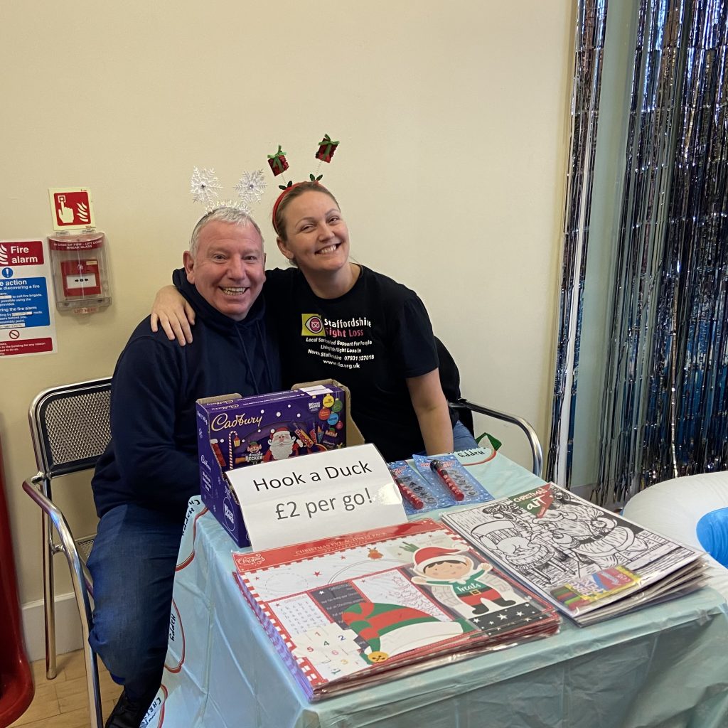 The image shows two smiling people sitting together at a table, likely at a festive event. The man on the left is wearing a dark hoodie and snowflake-themed headgear, while the woman on the right is wearing a black shirt with "Staffordshire Sight Loss" written on it, along with reindeer antlers headgear. They are sitting behind a small booth with a sign reading "Hook a Duck £2 per go!" There are festive-themed items on the table, including Cadbury chocolates and Christmas activity books. The background includes some tinsel decorations, adding to the festive atmosphere.