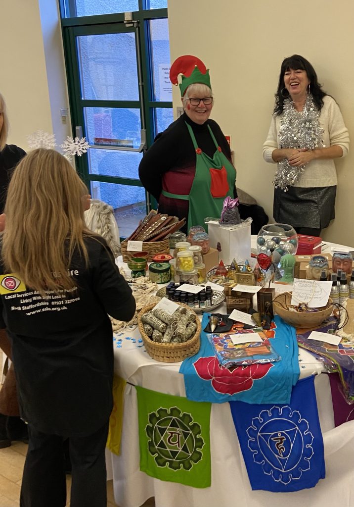 This image appears to show a festive event or a market stall. There are four women gathered around a table full of various handmade or decorative items. The woman in the middle is dressed in a Christmas elf costume, wearing a green and red apron with a matching elf hat. Another woman to the right is wearing a white sweater with silver tinsel around her neck, laughing and chatting. On the table are a variety of items, including incense sticks, candles, essential oils, sage bundles, and colorful flags with chakra symbols. The woman in the foreground, facing away from the camera, has a Staffordshire Sight Loss shirt. The scene is indoors, with a bright window in the background, likely part of a community gathering or holiday fair.