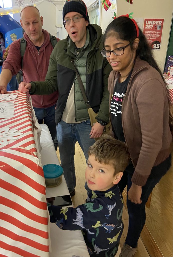 This image shows a group of people gathered around what appears to be a table covered with a red and white striped tablecloth, likely at some kind of event or gathering. There are four people visible in the image, A bald man in a red jacket on the left, A man in the middle wearing a black beanie hat, glasses, and a green and black jacket, A young woman with long dark hair and glasses wearing a brown sweater and a black t-shirt with text on it, and A young boy in the foreground wearing a blue shirt with dinosaur prints The man in the middle appears to have an surprised or excited expression, with his mouth open. The others are looking at something on the table, though we can't see what they're focused on. The setting seems to be indoors, possibly a community center or school. There are decorations visible in the background, including what looks like holiday-themed items. The atmosphere appears casual and engaged, with the group seemingly interested in whatever is on the table in front of them.