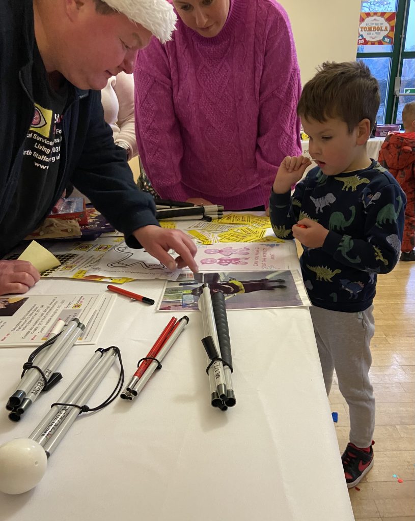 The image shows three people gathered around a table. On the left is an adult wearing a dark jacket with a white hat or cap. In the middle is an adult wearing a bright pink sweater. On the right is a young boy, probably around 4-6 years old, wearing a blue sweater with dinosaur prints. They appear to be engaged in some kind of activity or project on the table. There are various papers, markers, and what look like canes or mobility aids laid out on the table surface. The boy is watching intently as the adult on the left points to or works on something on the table. The setting seems to be indoors, possibly in a community centre or similar space. There's a sign visible in the background for the Tombola. The scene gives the impression of a learning or interactive activity, with the adults guiding or teaching the child about something spread out on the table.