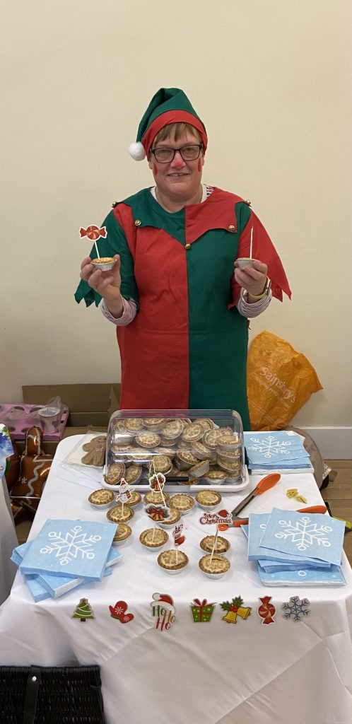 The image shows a person dressed as a Christmas elf standing behind a table with festive treats. They are wearing a red and green elf costume with a pointed hat that has a white pom-pom on top. The person is smiling and holding up small items in each hand. On the table in front of them is an assortment of Christmas-themed items: A large tray filled with what appear to be mince pies or small tarts Blue napkins with snowflake designs Small decorative items like gingerbread men, Christmas trees, and other holiday symbols along the front edge of the table Some larger decorations or signs, including one that says "Merry Christmas" The table is covered with a white cloth, and background is a plain beige wall. The overall scene suggests this person is working at or participating in a Christmas fair or event, offering festive treats to visitors.