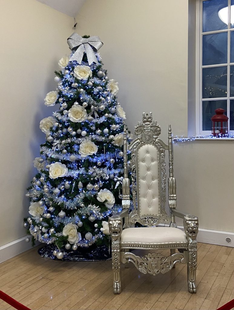 This image features a beautifully decorated Christmas tree standing next to a regal-looking chair. The tree is adorned with white paper roses, silver tinsel, and blue lights, creating a festive and elegant atmosphere. A large silver bow sits atop the tree. The chair is upholstered in white with a silver frame and lion paw feet, adding a touch of grandeur to the scene. A red lantern is visible in the background near a window, further enhancing the Christmas ambiance. The overall setting appears to be a cozy room with wooden floors.