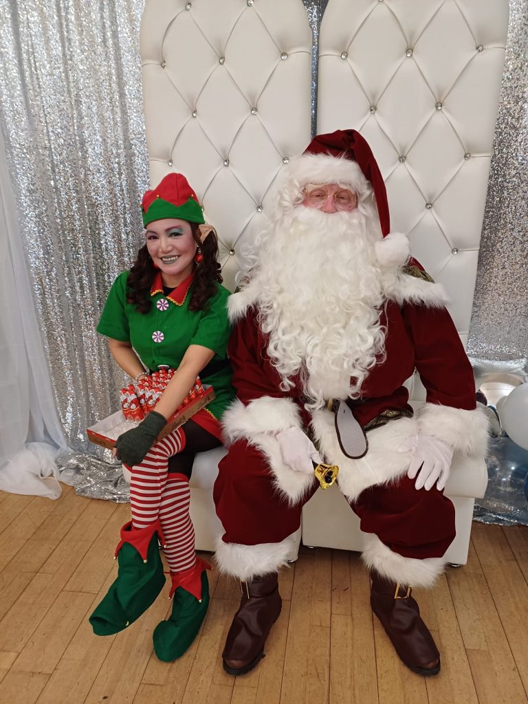 The image depicts a classic Christmas scene with Santa Claus and one of his elves. They are seated on a white, tufted chair or throne against a backdrop of shimmering silver curtains or decorations. Santa is dressed in his traditional red suit with white fur trim. He has a long, flowing white beard and is wearing glasses. His expression appears jolly and kind. Santa is holding a small bell in one hand. Seated next to Santa is a woman dressed as an elf. She is wearing a green shirt with candy cane buttons, red and white striped tights, and green elf shoes with curled toes. Her outfit is completed with a red and green elf hat with pointed ears attached. The elf has long dark hair and is smiling brightly. She is holding what looks like a tray of small red and white candy canes or similar treats. The setting appears to be a Christmas photo booth or display. The floor is hardwood, and the white tufted backdrop behind them has rhinestone or crystal embellishments. The overall atmosphere is festive and cheerful, capturing the spirit of the holiday season.