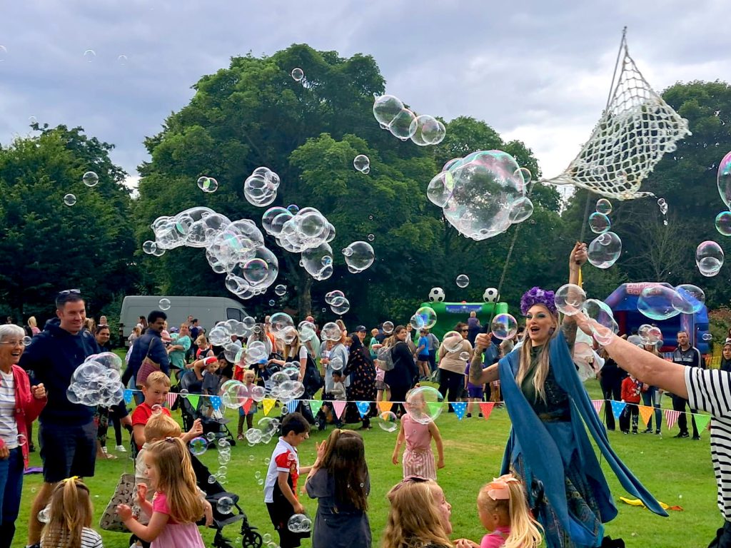 Image shows a large open green space in Burslem park, filled with people young and old enjoying themselves. In the foreground of the image is a lady dressed as a fairy in blue outfit with a purple headdress holding a large net from which bubbles are being produced. There is also a group off children looking at and playing with the bubbles which are floating around them and over their heads.