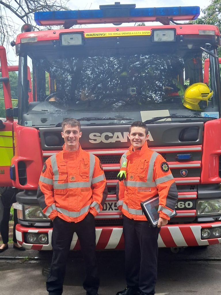 Image shows two fire personnel standing in their uniforms (navy trousers and bright orange high visibility coats) in front of the front their fire engine. The two men look as though they are enjoying themselves as they are smiling.