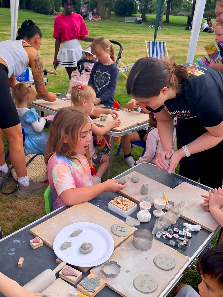 Image shows 2 tables to children enjoying a pottery craft. On the table are wooden trays with different pieces of clay which the children are playing with, there are also a number of tool – stamps, cutters etc. At one of the tables a volunteer is leaning over the table showing a young girl how to make a particular shape out of the clay