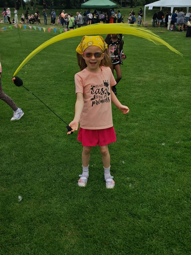 Image shows a young girl with a yellow headscarf, orange top and red shorts smiling while having a go at circus skills, the girl has a poi attached to her finger and is spinning it so its yellow tail forms a circle around her. Poi are round black balls with a colourful tail attached to the hand by a large piece of string that can be spun around to give a sort of juggling like illusion.