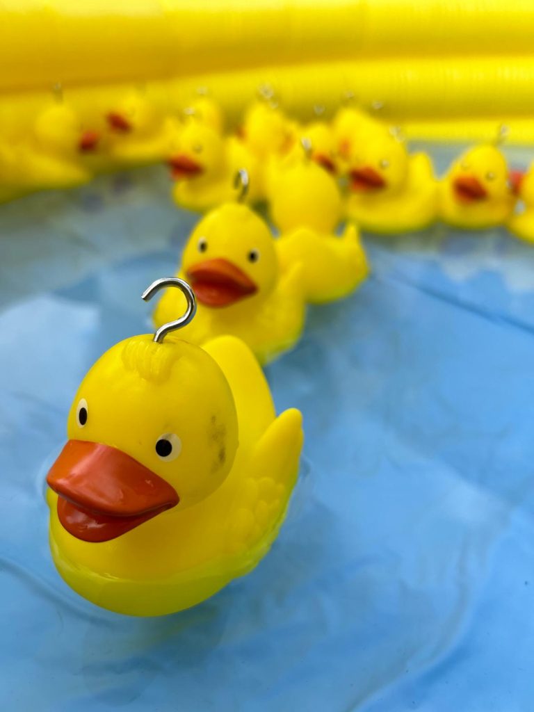 Image shows several yellow rubber ducks in a paddling pool of water set up for hook a duck. The bottom of the paddling pool is pale blue and the sides are yellow