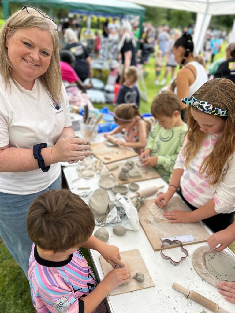 Image shows a very busy craft tent where Sara From HSL Pottery is looking at the camera smiling, stood around the table in front of her are a number of children all busy with their individual clay creations. There are lots of tools scattered about to help them with this, from cutters to rolling pins. The children are all concentrating hard on what they are doing.