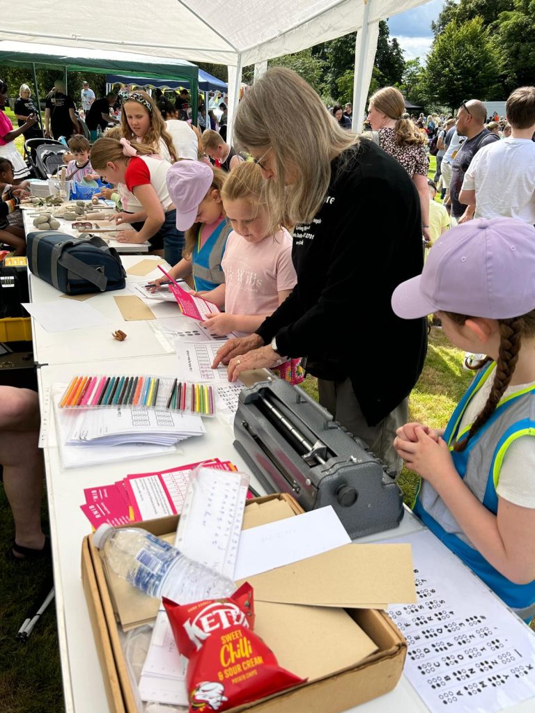 Image shows a table set out with items to help children learn a little basic braille. We can see copies of the braille alphabet, a perkins brailler, some colouring pencils and a box of braille paper. Jennifer is talking to one little girl about the braille dots she has coloured in, presumably to spell out her name while another girl listens on waiting for her turn.