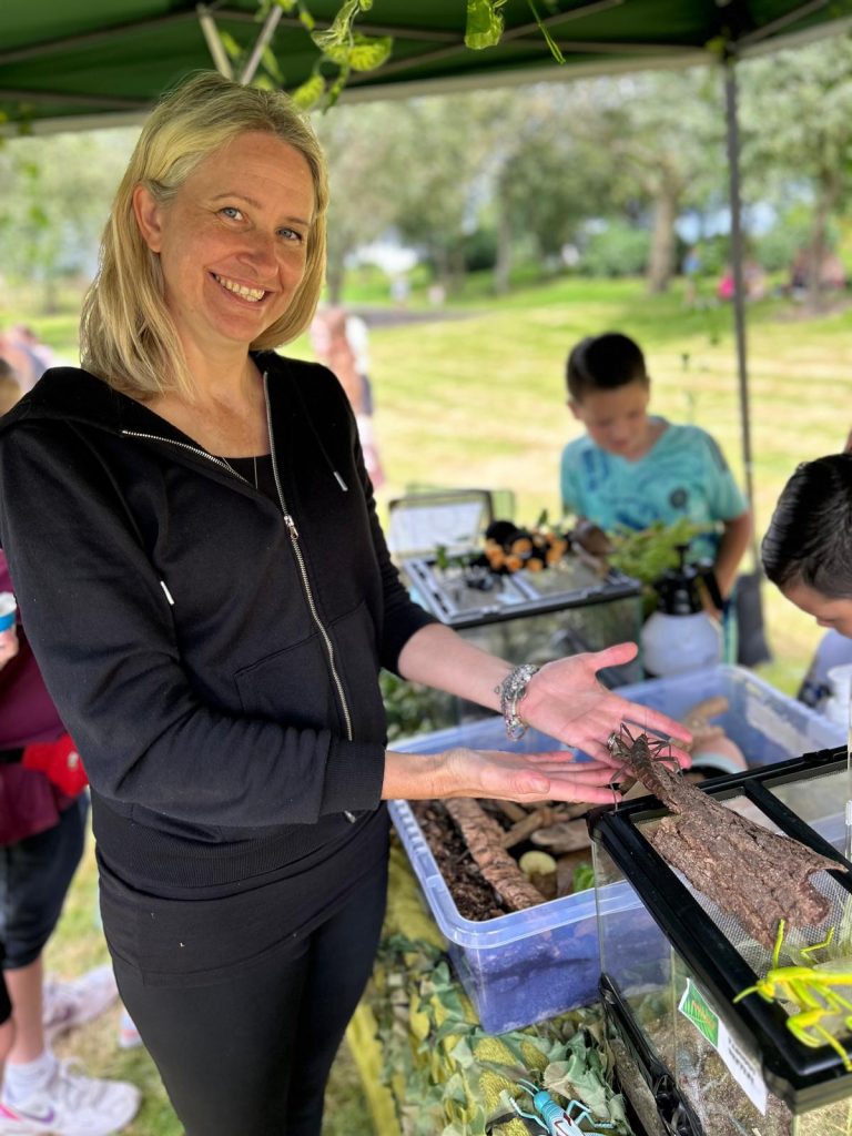 Image shows a blonde lady in black clothing smiling while holding a large bug in her hands. Her hands are held over a table with a few vivarium’s and plastic tubs on it which are acting as habitats for the creatures.