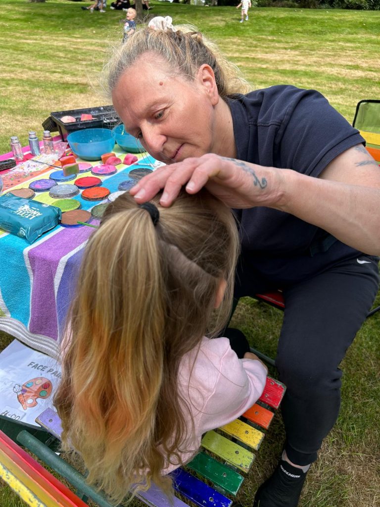 Image shows a lady painting the face of a little girl, we cannot see what image is being painted but the face painter is obviously concentrating hard. On the table next to the face painter there is a vast array of colours of face paint and sponges, water and brushes that are used to create the designs.