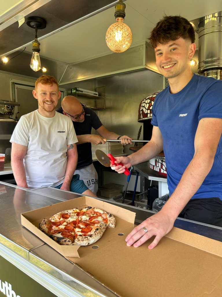 Image shows the inside of a food van with 2 staff members smiling at the camera with one of their pizzas on the counter in front of them. A third staff member is busy putting pizza into the pizza oven at the back of the food van.