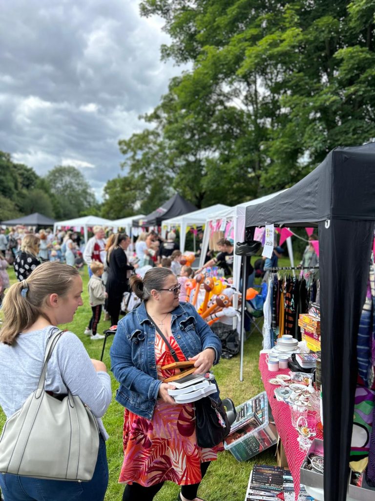 Image shows a row of marquees stretching off into the distance all with people gathered around the front of them, in the foreground is the second-hand items stall and the pocket money toys stall can be seen just behind it.