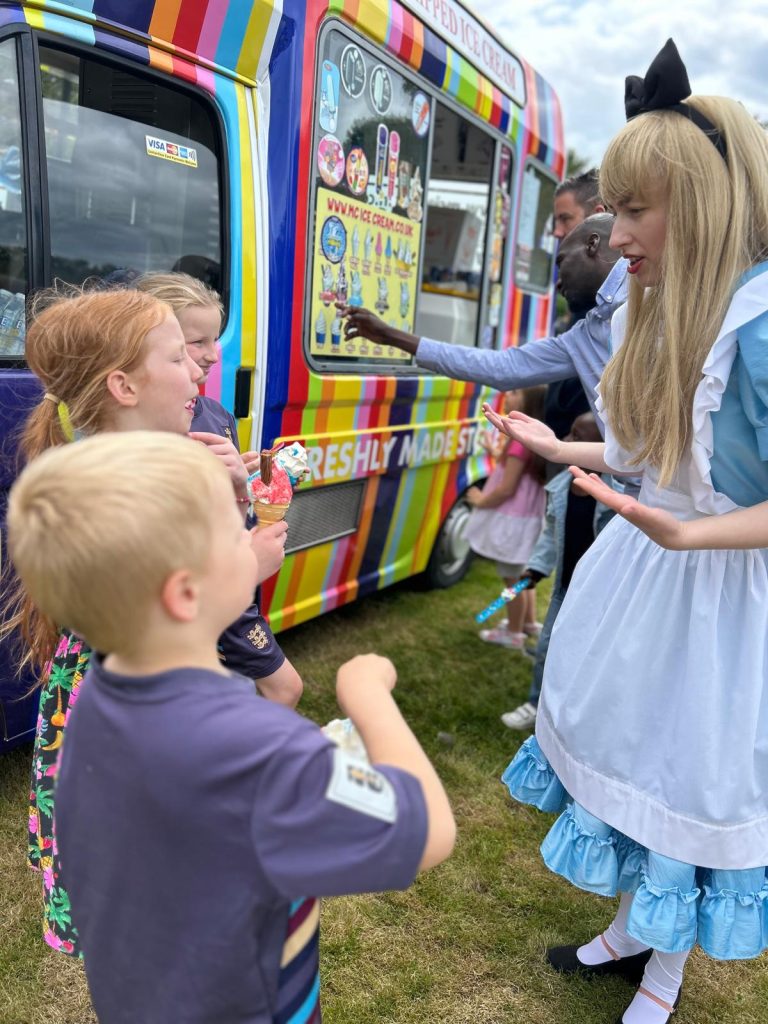 Image shows a lady dressed as Alice in wonderland talking to 3 children. The background of the picture is a colourful ice-cream van at which we can see children and adults choosing what ice cream they would like.